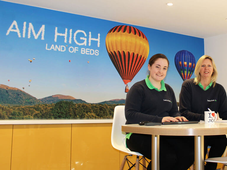 Two Land of Beds staff smiling, sitting in a room in the company's headquarters building. A logo on the wall behind them reads Aim High
