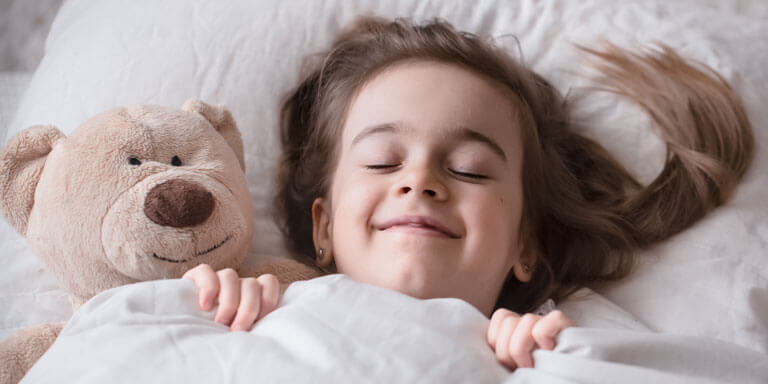 A young girl is fast asleep under the covers with a smile. Her teddy bear is nestled beside her on the pillow