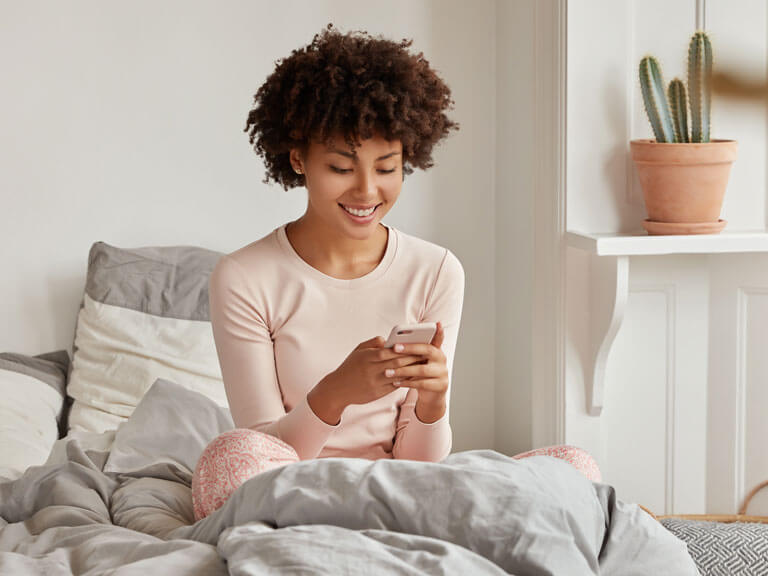 A woman is sitting on a bed, holding a phone. She appears to be looking at the phone screen and smiling