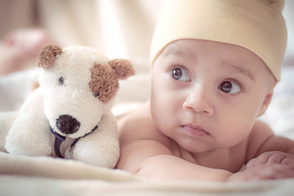 Baby wearing a yellow cap looking interested in something as a cute plush dog sits next to him