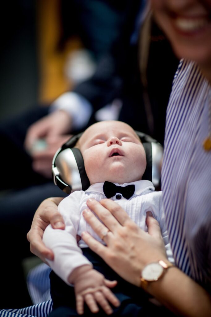 Baby sound asleep in his parent's arms as he wearing a tuxedo and headphones