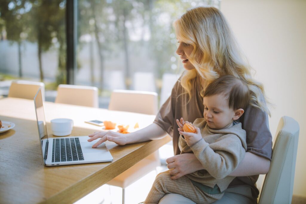 Woman working from home on laptop at her kitchen table as she holds her baby who is eating an orange