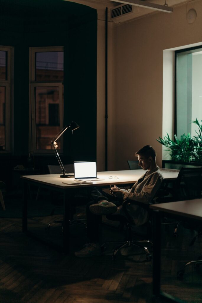 Man sat at a desk working from home with laptop and mobile illuminated by a lamp as he is working late