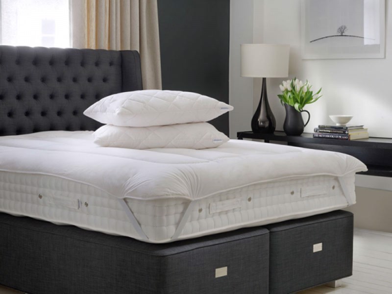 Grey bed base with a mattress laying on it featuring a mattress topper and some pillows