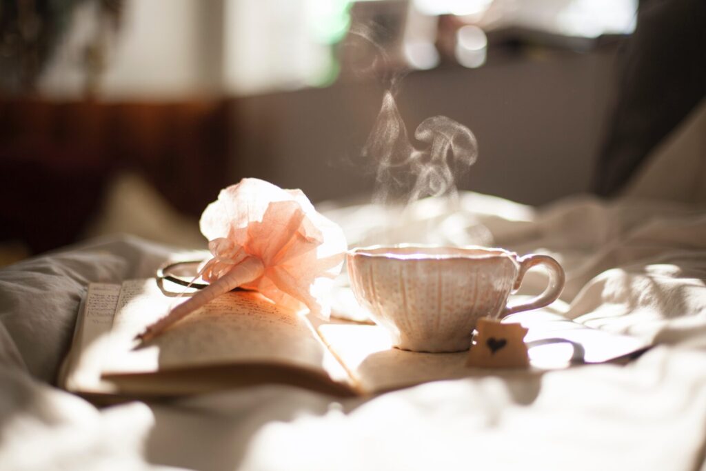 Steam rising from an elegant cup of tea sat on a bed next to an open notebook and a pen