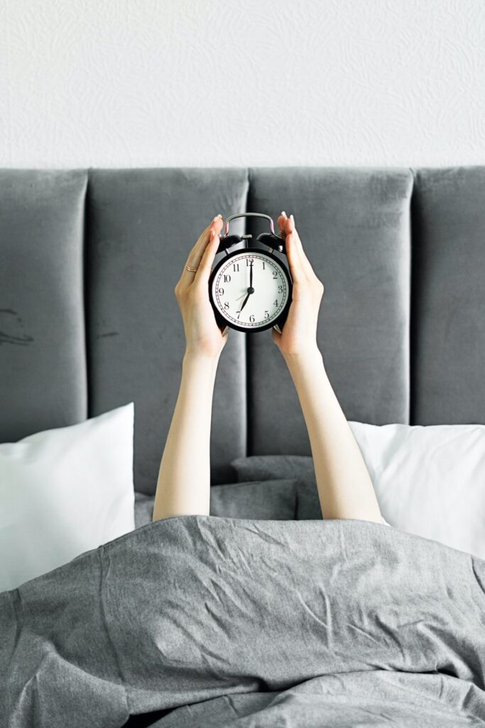 Woman hiding under duvet in bed but holding out an alarm clock