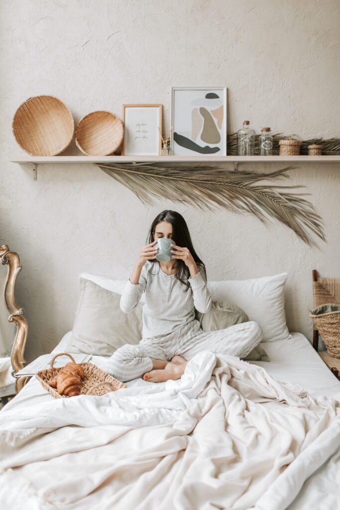 Woman enjoying hygge living on a bed drinking cocoa with a tray of croissants next to her and a beautifully decorated shelf above her