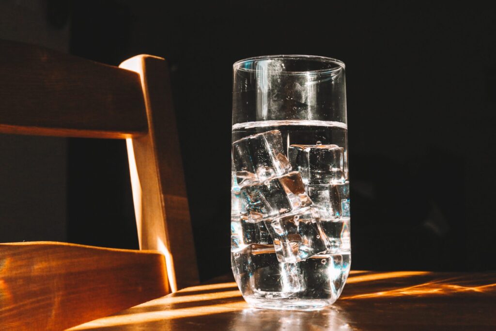 A glass of water stands proud atop a wooden table, a chair lurks near by. 