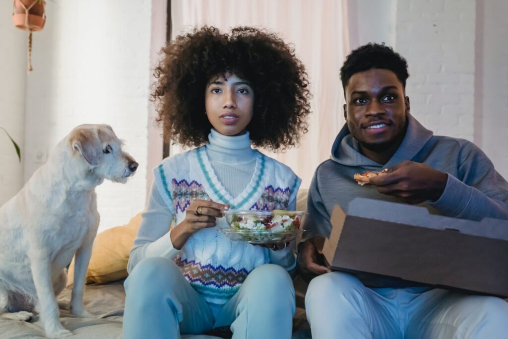Man and a woman sat on a bed eating as they watch something, possibly on a TV. A dog is on the bed eyeing up their food