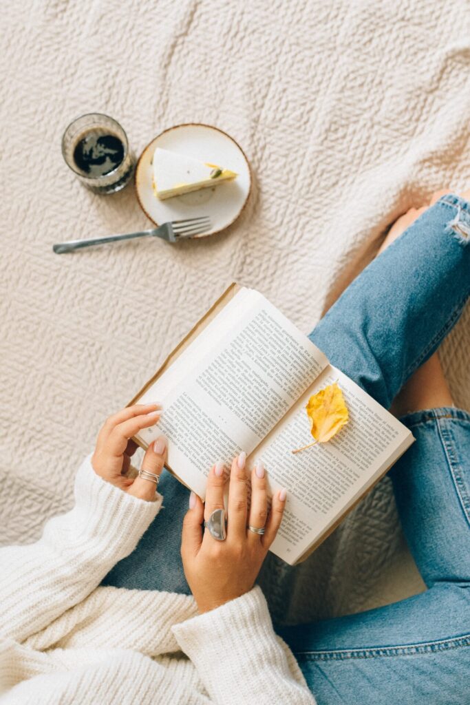 Woman enjoying hygge living on her bed by reading a book as a slice of cheese cake and a drink wait by her side