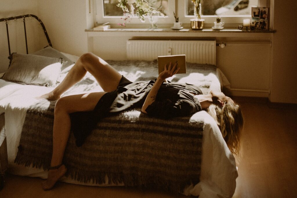 A woman lying on her bed while clutching a book but looking out the window