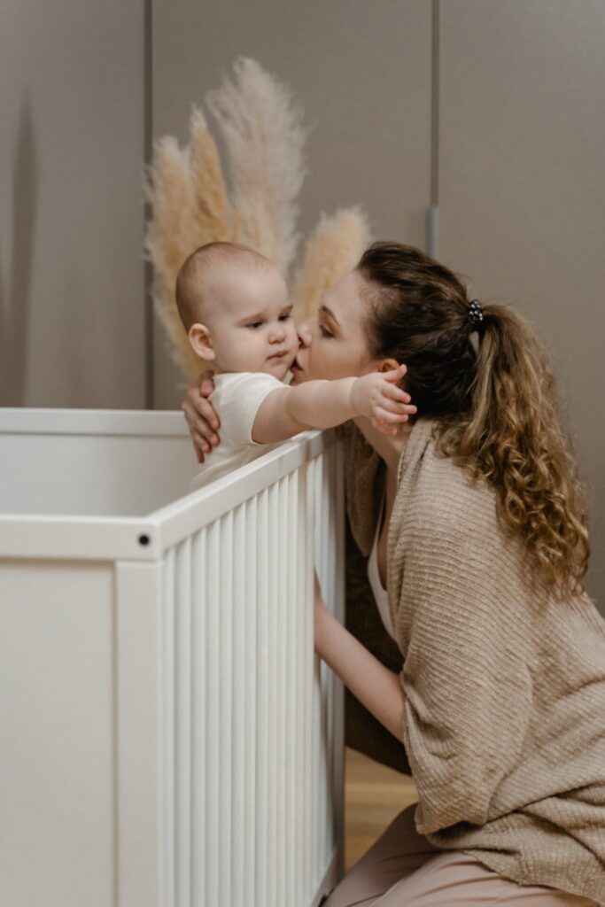Mother kissing her baby who is stood up in the crib