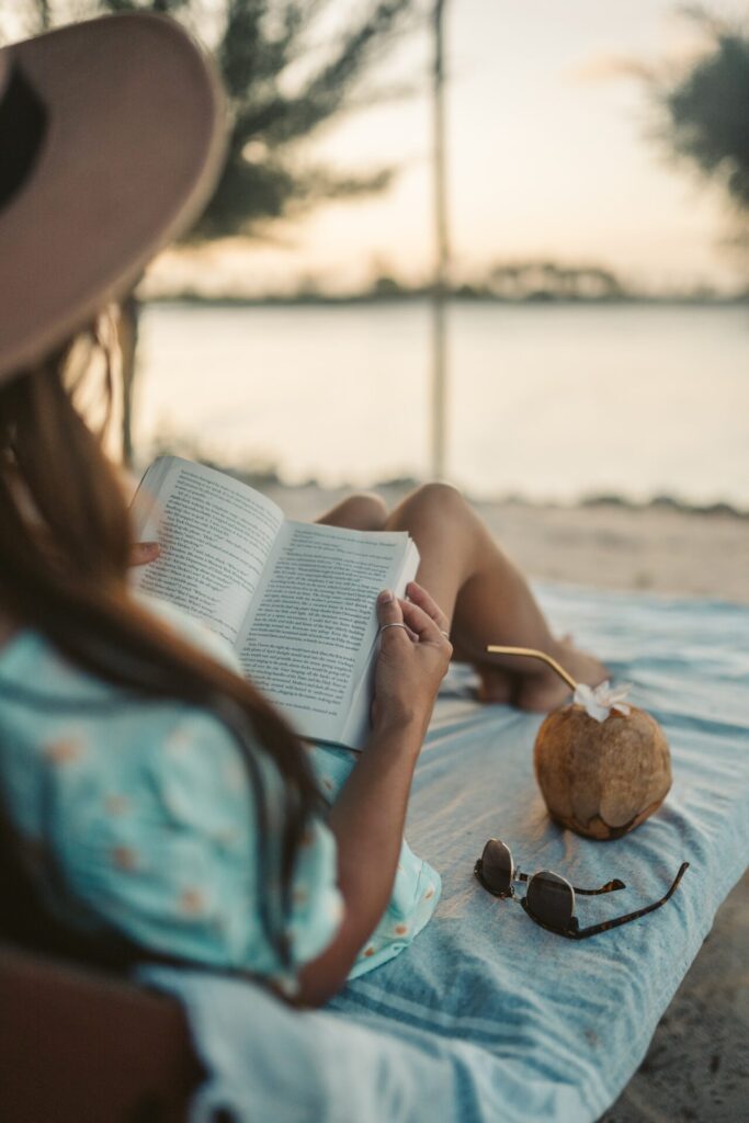 Woman sat on a beach reading a book while a coconut with a straw sits next to her.