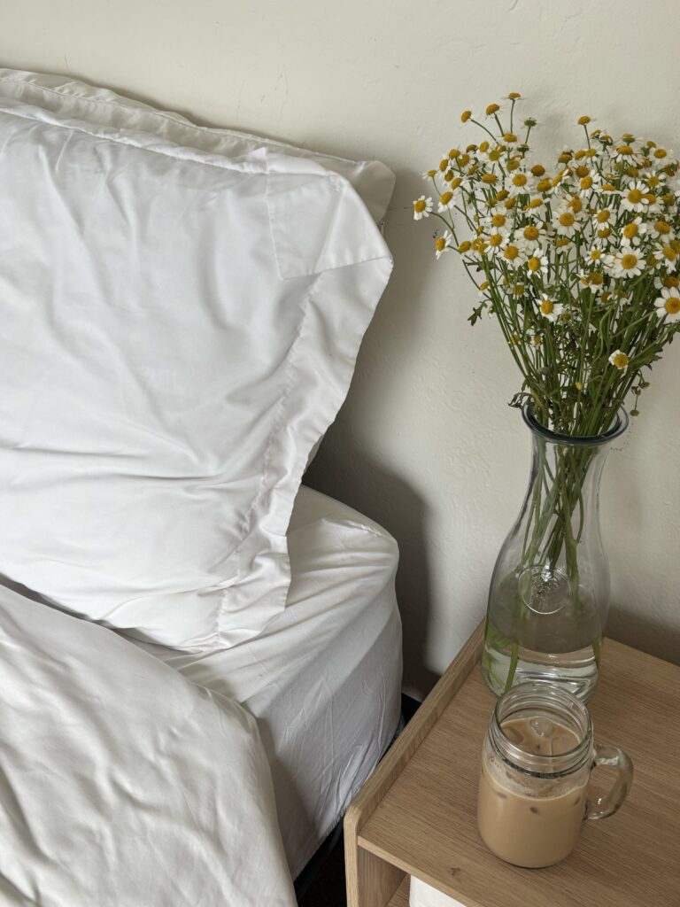 White bedding beside a wooden bedside table with fresh flowers and an iced coffee